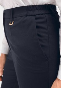 Marineblauwe tapered broek met elastische tailleband, gouden vierkante accenten en zichtbare zijzakken. De stof is glad met een minimale textuur.