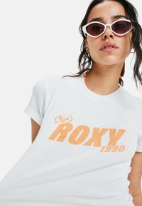 Witte katoenen t-shirt met korte mouwen en oranje "ROXY 1990" grafische print, gecombineerd met witte hoekige zonnebril.