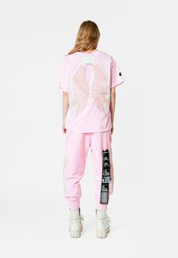 Camiseta rosa con diseño de alas en la parte posterior, combinada con pantalones de chándal rosas a juego que tienen una etiqueta gráfica lateral y botas blancas.
