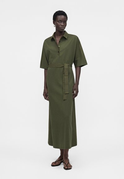 Γυναίκα που φοράει μεσαίο φόρεμα ελιάς με γιακά και μανίκια έως τον αγκώνα, με ζώνη στη μέση και μαύρα σανδάλια τύπου σαγιονάρα, στέκεται σε απλό φόντο.