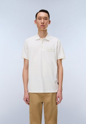 Young man standing against plain background wearing a white short-sleeve polo shirt and light brown pants, facing forward with a neutral expression.