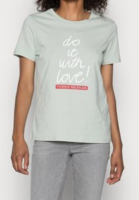 Camisa de manga corta verde claro con el texto "¡hazlo con amor!" y el logo rojo de "Tommy Hilfiger" que lleva puesta una persona con jeans grises.
