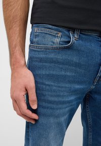 Ciemnoniebieskie dżinsy mają klasyczny krój z pięcioma kieszeniami, lekkimi przetarciami i kontrastującym pomarańczowym szwem. Ręka spoczywa na udzie.