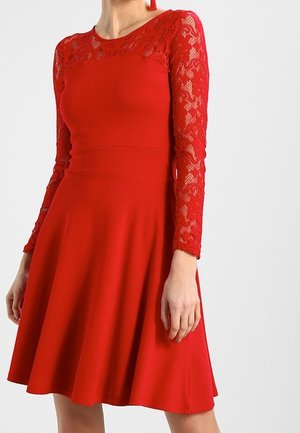 Vrouw draagt een rode jurk met lange kanten mouwen en een gevormd lijfje, uitlopend in een rok tot op de knie.