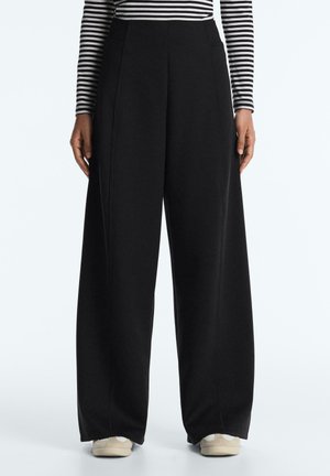 Személy magas derekú, bőszárú fekete nadrágot visel oldalzsebekkel, fekete-fehér csíkos hosszú ujjú felsővel és fehér sportcipővel.