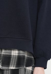 Sweat-shirt bleu marine avec des poignets côtelés, associé à un pantalon à motifs carreaux noirs et blancs, montrant les détails de texture et de tissu.