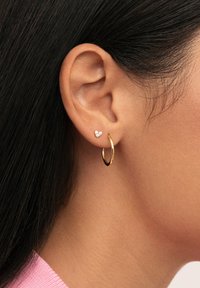 Pendiente de aro dorado acompañado de un pequeño pendiente en forma de corazón que presenta tres gemas transparentes. Superficie lisa, diseño moderno.