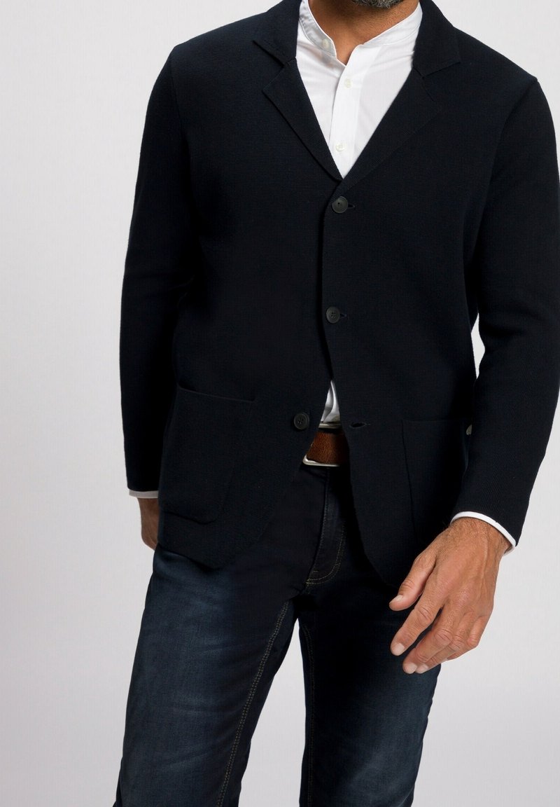 Blazer negro de punto con un corte entallado, cierre de un botón y dos bolsillos frontales, combinado con jeans oscuros y una camisa blanca.