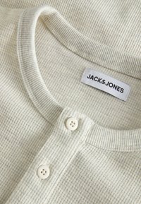 Beige waffle-textured knit shirt featuring a round neckline, button placket with two white buttons, and a label displaying "JACK & JONES."