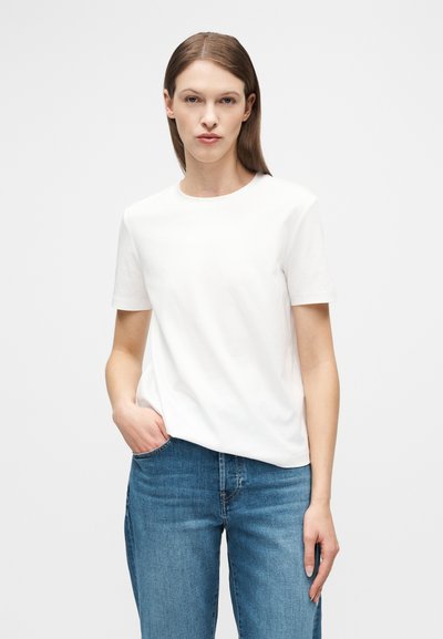 Γυναίκα με ίσια καστανά μαλλιά, που φοράει απλό λευκό κοντομάνικο μπλουζάκι και μπλε τζιν, όρθια με το ένα χέρι στην τσέπη.