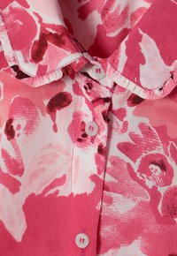 Lyserød bluse med blomsterprint, der har krave, knaplukning og en tekstureret stof med varierende nuancer af lyserød og rød.