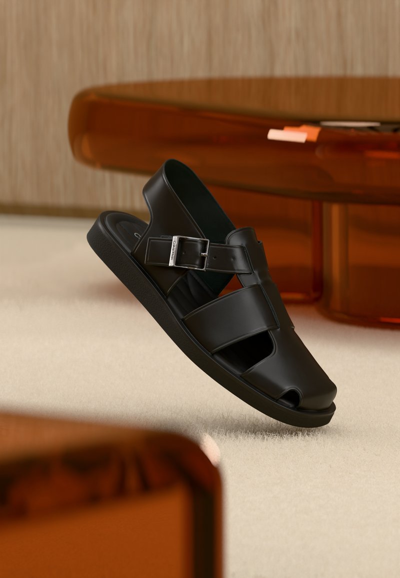 Μαύρο δερμάτινο σανδάλι με λουράκι και πόρπη, σχεδιασμός ανοιχτών δακτύλων, παρουσιάζεται σε μπεζ χαλί με θολό καφέ φόντο επίπλων.