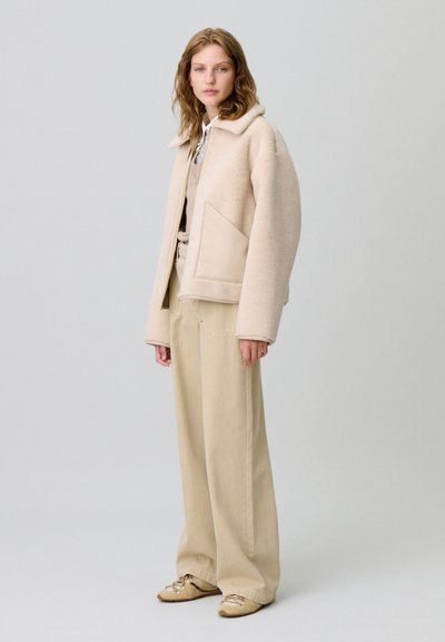 Giacca in shearling beige con colletto, una grande tasca laterale, abbinata a pantaloni larghi color miele e calzature chiare, su uno sfondo neutro.
