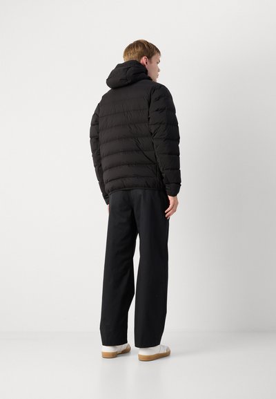 Doudoune noire avec capuche, présentant des coutures horizontales et une texture brillante, associée à un pantalon noir ample et des chaussures blanches.