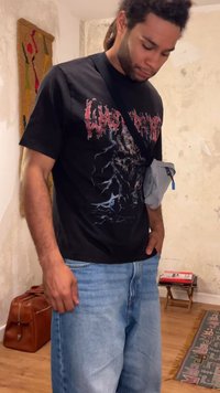 Άντρας με dreadlocks που φοράει ένα μαύρο γραφικό t-shirt και ανοιχτό μπλε τζιν στέκεται σε εσωτερικό χώρο δίπλα σε μια καφέ τσάντα και ένα μικρό τραπέζι με βιβλία.