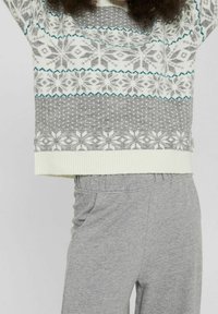 Person, die einen weißen und grau gemusterten Pullover mit Schneeflockenmotiven und graue Jogginghose trägt, während sie die Arme über den Kopf hebt.