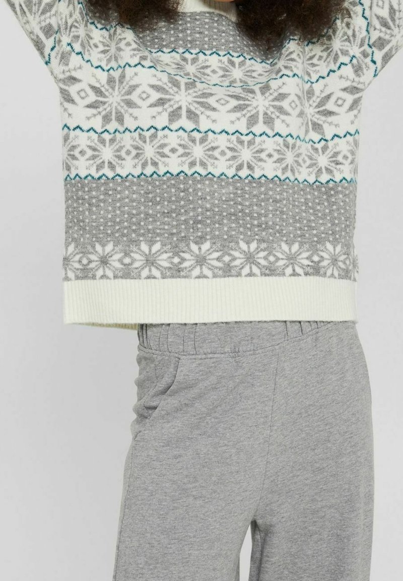 Person, die einen weißen und grau gemusterten Pullover mit Schneeflockenmotiven und graue Jogginghose trägt, während sie die Arme über den Kopf hebt.