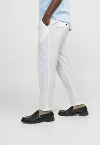 Man som bär vita byxor, ljusblå skjorta, beige strumpor och svarta loafers, stående med händerna i fickorna mot en enfärgad bakgrund.