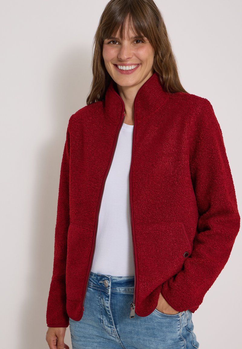 Femme souriante portant une veste en polaire rouge par-dessus une chemise blanche et un jean bleu, avec une main dans la poche.
