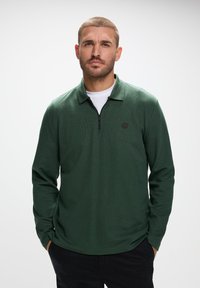 Ciemnozielona polówka z długim rękawem, z ćwierć zamkiem, płaskim ko�łnierzykiem i małym czarnym logo na klatce piersiowej, wykonana z teksturowanego materiału dzianinowego.