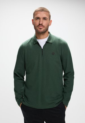 Mørkegrøn langærmet polo shirt med en kvart lynlås, flad krave og et lille sort logo på brystet, lavet af et struktureret strikmateriale.