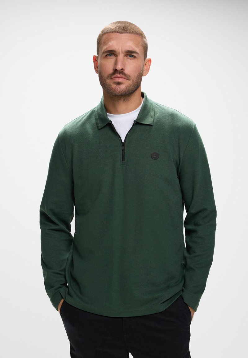 Ciemnozielona polówka z długim rękawem, z ćwierć zamkiem, płaskim kołnierzykiem i małym czarnym logo na klatce piersiowej, wykonana z teksturowanego materiału dzianinowego.