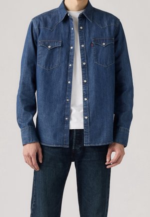 Homme portant une chemise en denim bleu foncé avec des boutons-pression sur un t-shirt blanc et un jean bleu foncé assorti, debout devant un fond gris.