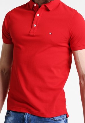 Homme portant un polo rouge ajusté à manches courtes avec deux boutons blancs, associé à un jean bleu en denim.
