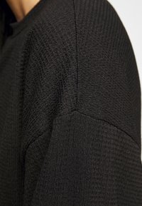 Černý pletený svetr s texturou zaměřený na rukáv; má ribovaný vzor, zaoblené ramenní švy a měkký, mírně vyvýšený povrch látky.