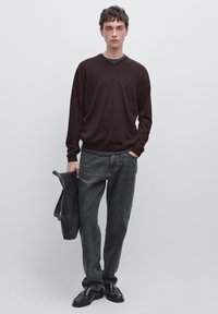 Dunkelbrauner V-Ausschnitt-Pullover, graue Denim-Jeans, schwarze Schuhe, hält eine schwarze Tasche. Einfaches Design, normale Passform, glatte Textur, minimale Details.