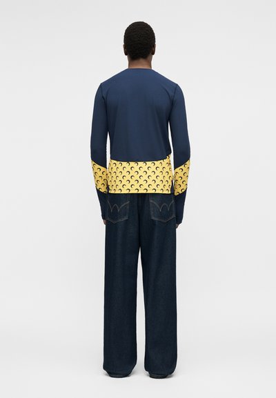 Άνδρας που φοράει μπλε μακρυμάνικη μπλούζα με κίτρινα μανσέτες και τελείωμα με σχέδιο μισοφέγγαρου, σε συνδυασμό με χαλαρά σκουρόχρωμα μπλε τζιν, γυρισμένος πλάτη.