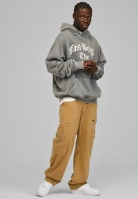 Grå oversized hoodie med hvid tekst, parret med tan cargo bukser med lommer, og hvide sneakers. Glat stof og afslappet pasform.