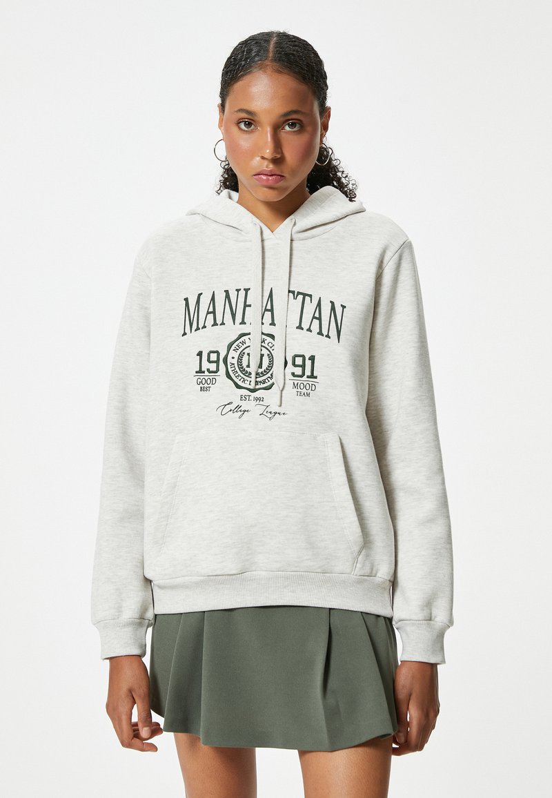 Sudadera gris claro hecha de una tela suave, con un bolsillo tipo canguro, capucha con cordón y un gráfico de texto negro que dice "MANHATTAN".