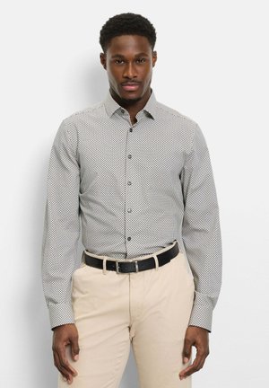 Mann trägt ein beige gemustertes Hemd mit Knöpfen, das in helle beige Hosen mit schwarzem Gürtel gesteckt ist, steht vor einem weißen Hintergrund.