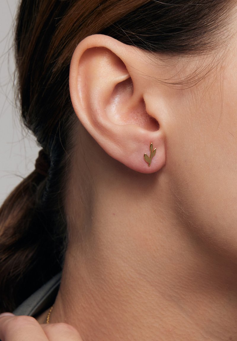 Kolczyk w kształcie liścia z złotego liścia na ludzkiej uchu, z gładkim wykończeniem i minimalistycznym designem. W pobliskim obszarze widoczna jest tekstura skóry.