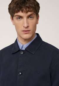 Donker marineblauwe jas met knopen aan de voorkant, voorzien van een brede kraag en een subtiel logo op de borst. Daaronder zit een lichtblauw patroonjasje.