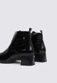 Botines de charol negro con tacón cuadrado, puntera redondeada, tiradores y un sutil detalle de costura en los laterales.