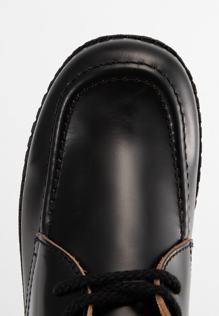 Chaussure en cuir noir verni avec bout rond, couture détaillée et lacets noirs sur fond blanc.