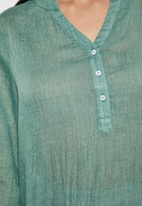 Camicia di lino verde chiaro con scollo a V e tre bottoni. Presenta un tessuto testurizzato e un orlo a punta sul colletto.