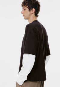 Schwarzes kurzärmeliges T-Shirt, das über einem weißen langärmeligen Hemd getragen wird, mit einem lockeren Schnitt und Rundhalsausschnitt, von der Seite gezeigt.
