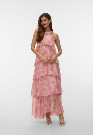 Donna che indossa un vestito lungo rosa a balze con motivi floreali, collo halter e dettaglio di rosa, in piedi e sorridente davanti a uno sfondo semplice.