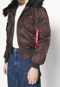 Muž v hnědé bomber jacket s černým kožešinovým límcem, bílém tričku a modrých džínách, s červenou visačkou s nápisem "Odstranit před letem" na kapsičce rukávu.