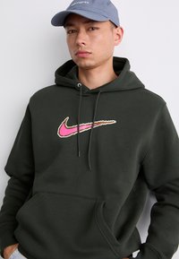 Mørkegrøn hættetrøje med et stort pink og hvidt Nike-logo, snoretræk og en frontlomme. Båret med en lyseblå kasket.
