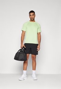Green short-sleeve athletic shirt, black shorts, white sneakers, carrying a black duffel bag. Simple design, lightweight material, minimal accents.