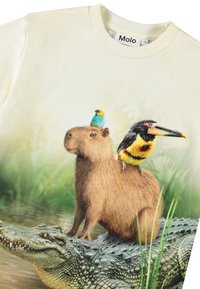 Vaaleanbeige T-paita, jossa on kapibara sinisen ja keltaisen linnun päällä, sekä musta, keltainen ja punainen lintu, vihreää taustaa vasten.