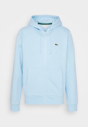 Sweat à capuche bleu clair avec fermeture éclair, cordons, poches avant, et petit logo crocodile vert Lacoste sur le côté gauche de la poitrine.