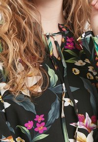 Svart blus med livfullt blommönster som har gröna blad och rosa, vita och gula blommor. Binder i halsen, mjuk tygtextur.