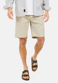 Beige shorts lavet af letvægtigt stof, med lige snit og sidelommer, parret med sorte sandaler med to stropper, der fremhæver en naturlig tekstur.