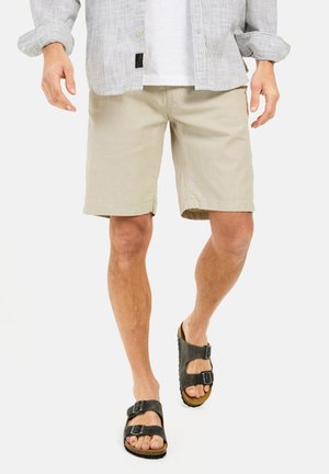 Beige shorts lavet af letvægtigt stof, med lige snit og sidelommer, parret med sorte sandaler med to stropper, der fremhæver en naturlig tekstur.