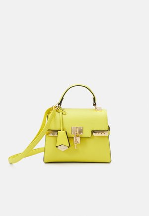 Jasnożółta torebka z uchwytem górnym, odpinanym paskiem na ramię, złotą kłódką z zawieszką w kształcie klucza i dekoracyjną metką na białym tle.